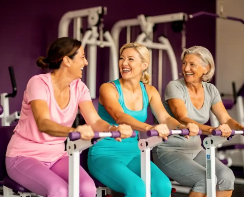 Drie vrouwen van middelbare leeftijd lachen samen tijdens het sporten op hydraulische fitnessapparaten in een paarse damesgymnastiek.