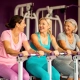 Drie vrouwen van middelbare leeftijd lachen samen tijdens het sporten op hydraulische fitnessapparaten in een paarse damesgymnastiek.