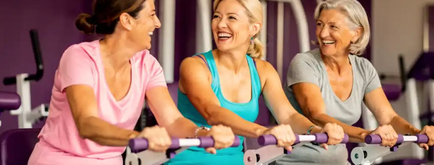 Drie vrouwen van middelbare leeftijd lachen samen tijdens het sporten op hydraulische fitnessapparaten in een paarse damesgymnastiek.