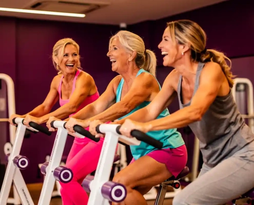 Drie vrouwen van middelbare leeftijd lachen samen tijdens een training op hydraulische fitnessapparaten in een moderne damesgymnastiek met paarse muren.