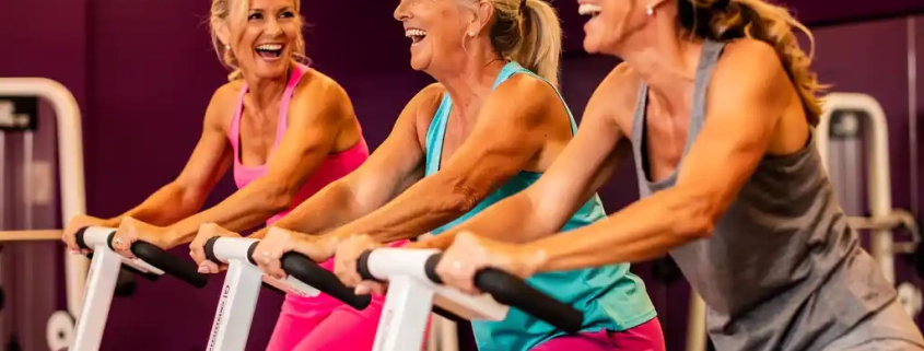 Drie vrouwen van middelbare leeftijd lachen samen tijdens een training op hydraulische fitnessapparaten in een moderne damesgymnastiek met paarse muren.