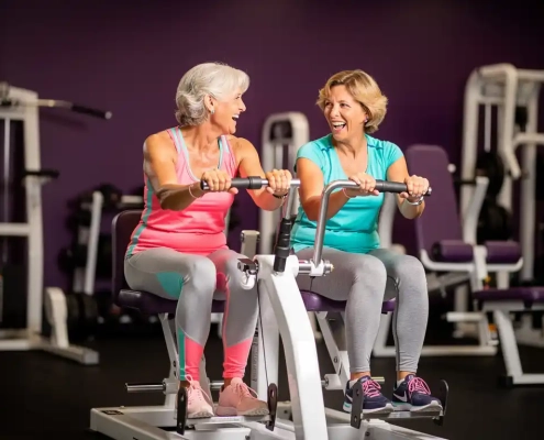 Twee vrouwen van in de vijftig lachen samen tijdens een work-out op hydraulische fitnessapparaten in een vrouwengymnastiek met paarse muren.
