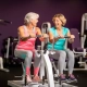 Twee vrouwen van in de vijftig lachen samen tijdens een work-out op hydraulische fitnessapparaten in een vrouwengymnastiek met paarse muren.