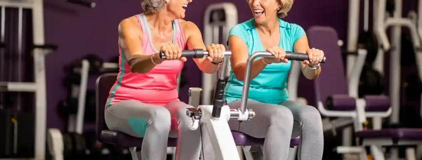 Twee vrouwen van in de vijftig lachen samen tijdens een work-out op hydraulische fitnessapparaten in een vrouwengymnastiek met paarse muren.