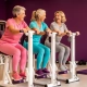 Drie vrouwen van 50-60 jaar lachen en sporten samen op hydraulische fitnessapparaten in een paarse damesfitnessruimte.
