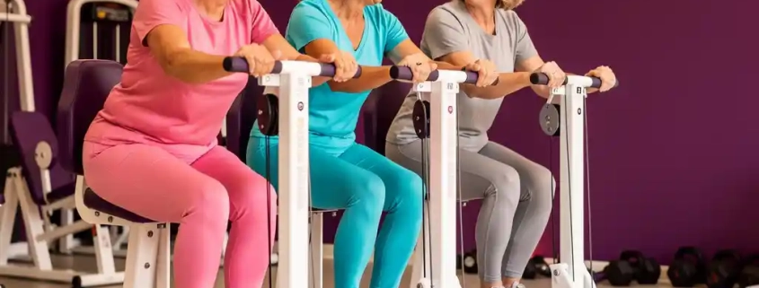 Drie vrouwen van 50-60 jaar lachen en sporten samen op hydraulische fitnessapparaten in een paarse damesfitnessruimte.