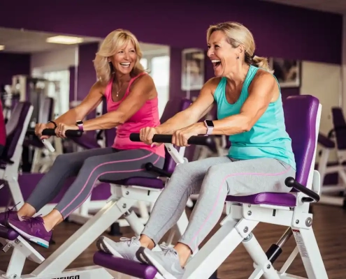 Twee vrouwen lachen samen tijdens een training op hydraulische fitnessapparaten in een vrouwenfitnessclub met paarse muren.