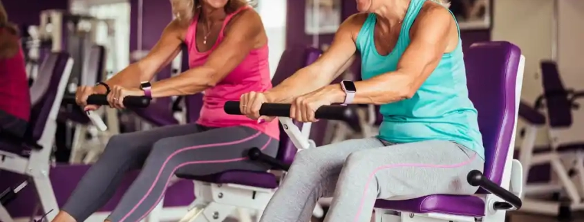 Twee vrouwen lachen samen tijdens een training op hydraulische fitnessapparaten in een vrouwenfitnessclub met paarse muren.