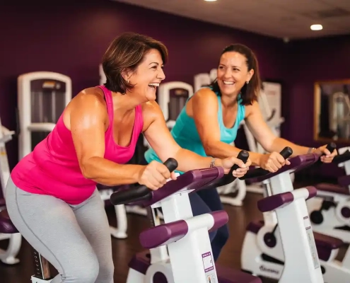 Twee vrouwen lachen samen tijdens een krachttraining op hydraulische fitnessapparaten in een levendige vrouwenfitnessclub met paarse muren.