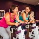 Twee vrouwen lachen samen tijdens een krachttraining op hydraulische fitnessapparaten in een levendige vrouwenfitnessclub met paarse muren.