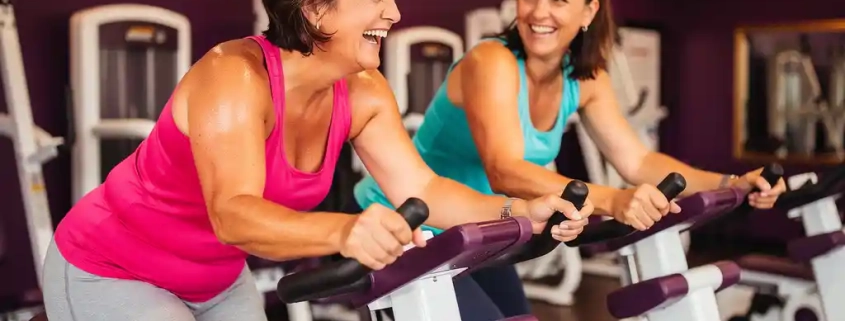 Twee vrouwen lachen samen tijdens een krachttraining op hydraulische fitnessapparaten in een levendige vrouwenfitnessclub met paarse muren.