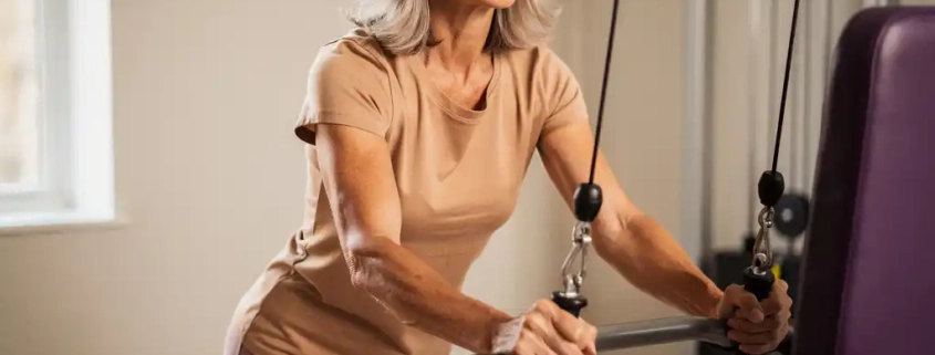 Vrouw van middelbare leeftijd traint gefocust op hydraulische weerstandsmachine in lichte fitnessstudio.