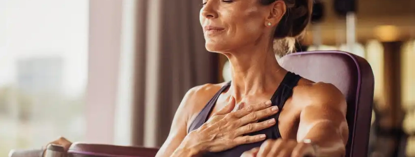 Vrouw van middelbare leeftijd pauzeert tijdens krachttraining op hydraulisch fitnessapparaat, hand op borst, ogen gesloten.