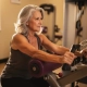 Vrouw van middelbare leeftijd met zilvergrijs haar traint rustig op hydraulisch fitnessmachine in Curves-studio met warm ochtendlicht.