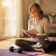 Vrouw van middelbare leeftijd met grijs haar schrijft in wellness-dagboek op yogamat bij zonnig raam, kleine halter naast haar.